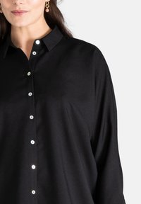 Camicia nera a bottone realizzata in tessuto morbido, con colletto classico, bottoni simili a perle e una vestibilità rilassata.