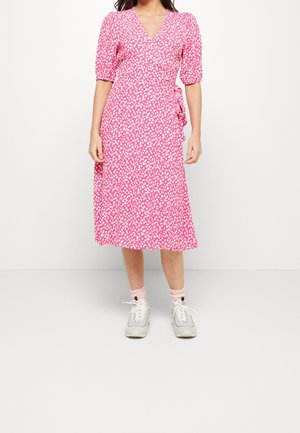 Vrouw draagt een roze midi wikkeljurk met witte abstracte vlekken, opgeblazen mouwen, gecombineerd met lichtroze sokken en witte sneakers.