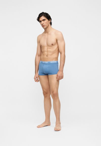 Άνδρας χωρίς πουκάμισο, όρθιος ξυπόλητος, φορώντας μπλε εσώρουχα boxer Moschino σε απλό λευκό φόντο.