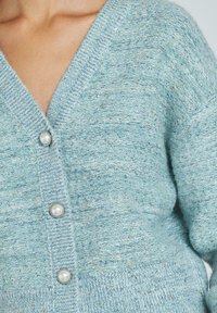 Cardigan bleu deschis cu decolteu în V, model tricotat texturat și accente cu nasturi din perlă. Materialul pare moale, cu o strălucire subtilă.