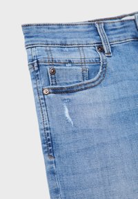 Jeans de denim azul claro con detalles desgastados, cinco bolsillos y acentos de metal cobre. Textura suave con líneas de costura visibles.