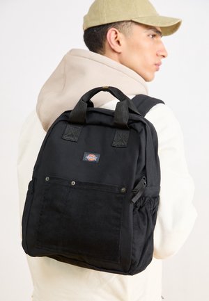 Joven con gorra beige y sudadera con capucha, llevando una mochila negra con bolsillo delantero y bolsillos laterales de malla, mirando hacia la derecha sobre un fondo blanco liso.