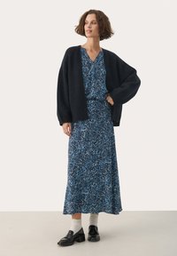 Cardigan nero in maglia sopra un vestito floreale blu. Il vestito è lungo con una silhouette fluida. Si indossano mocassini neri e calze grigie chiare.