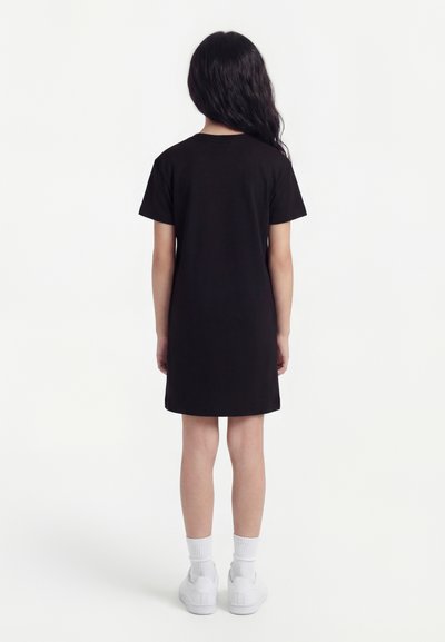 Κορίτσι που στέκεται με την πλάτη γυρισμένη φορώντας μαύρο φόρεμα με κοντά μανίκια, λευκές κάλτσες και λευκά αθλητικά παπούτσια μπροστά σε απλό λευκό φόντο.