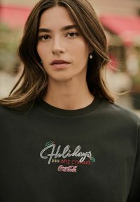 Dark green sweatshirt featuring embroidered text "Holidays Are Coming" with floral accents and a Coca-Cola logo. Round neckline and soft texture.