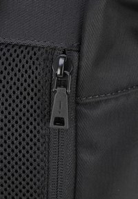 Zaino in tessuto nero con pannello in rete, dotato di una cerniera liscia e di un tassello in plastica. Il materiale strutturato aggiunge contrasto al design.