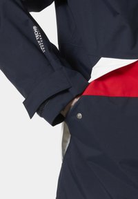Marineblauwe waterdichte jas met een contrasterende rode accent, verstelbare manchetten en een snap closure pocket detail.