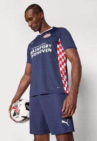 Marinblå idrottströja med röd och vit rutmönstrad sidopanel och vit text. Kombinerad med marinblå shorts. Spelaren håller en svart och vit fotboll.