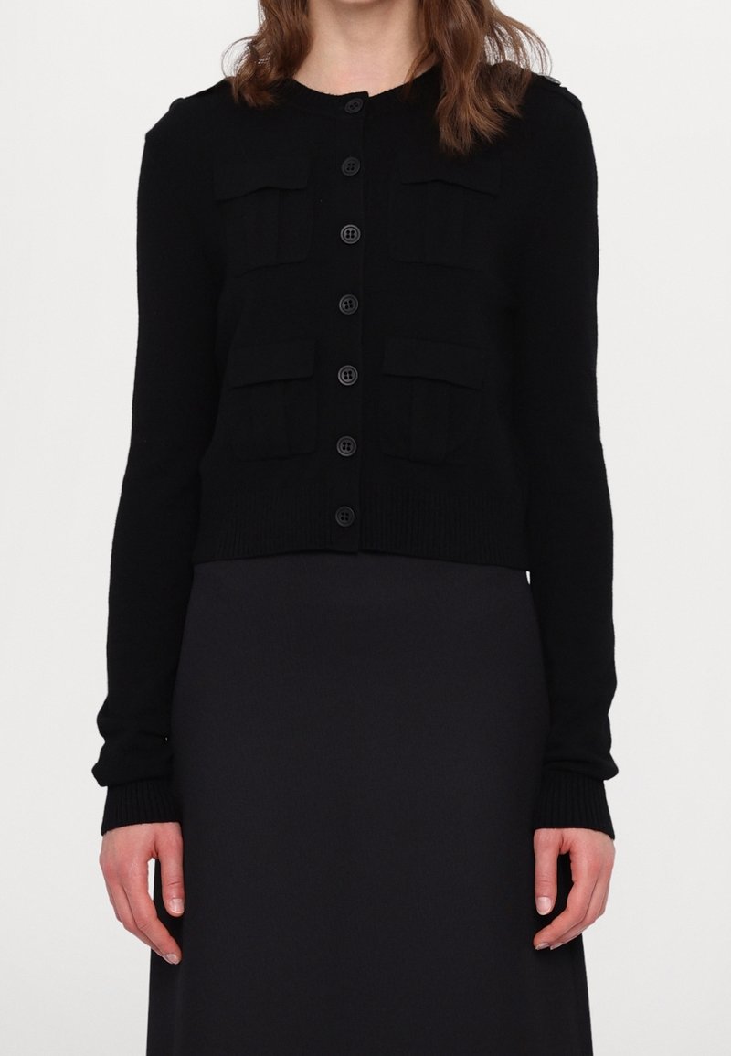 Femme portant un cardigan noir à manches longues boutonné avec quatre poches devant sur une jupe noire.