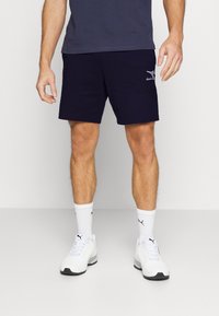 Shorts de sport navy avec poches latérales, taille élastique et logo brodé. Associés à des baskets blanches et des chaussettes blanches côtelées.