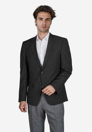 Schwarze Sakkajacke mit einem Zweiknopfverschluss, Reverskragen und einer einzigen Brusttasche, kombiniert mit grauen Karohosen. Darunter ein weißes Hemd.