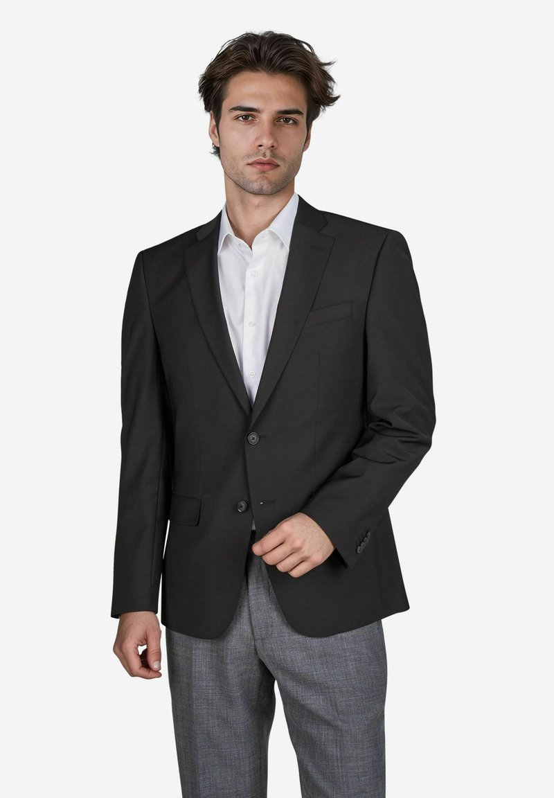 Schwarze Sakkajacke mit einem Zweiknopfverschluss, Reverskragen und einer einzigen Brusttasche, kombiniert mit grauen Karohosen. Darunter ein weißes Hemd.