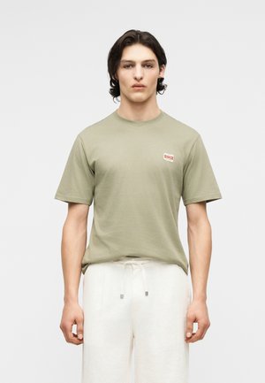 Νέος άνδρας με μαύρα μαλλιά μέχρι τους ώμους, που φοράει ένα πράσινο χακί μπλουζάκι και άσπρο παντελόνι με κορδόνι, στέκεται μπροστά σε ένα απλό λευκό φόντο.