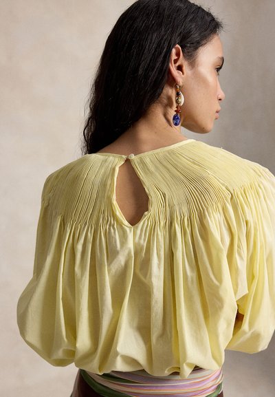 Mujer de perfil con cabello largo y oscuro, vistiendo una blusa plisada de color amarillo pálido con una abertura en la parte trasera abotonada y un cinturón de rayas coloridas.