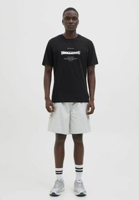 Sort bomulds T-shirt med et hvidt grafisk print på forsiden, parret med grå shorts, hvide sokker og grå sneakers. Enkelt design og afslappet pasform.