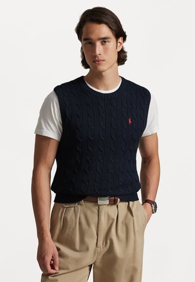 Chaleco de punto trenzado azul marino con dobladillo acanalado, usado sobre una camiseta blanca. Presenta un pequeño logotipo rojo en el pecho y pantalones beige.