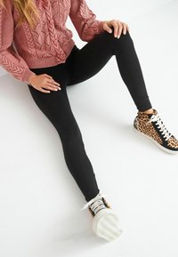 Pullover in een stoffige roos met kabelbreipatronen, gecombineerd met zwarte leggings en sneakers met luipaardprint, zittend tegen een witte achtergrond.