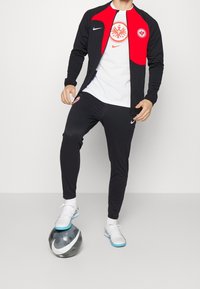 Chándal Nike en negro con detalles en rojo, que presenta un logo del club en el pecho y en el muslo, combinado con una camiseta blanca y calzado con suela azul.