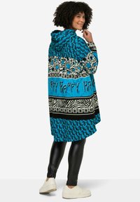 Blauwe gemusterde gevoerde jas met zwarte en witte accenten, verschillende texturen en ontwerpen, gecombineerd met zwarte leggings en witte schoenen.