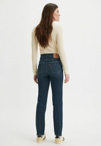 Högmidjade mörkblå denimjeans med en broderad Levis-logotyp på baksidan, slim fit och subtil blekning. Bärs med en beige topp.