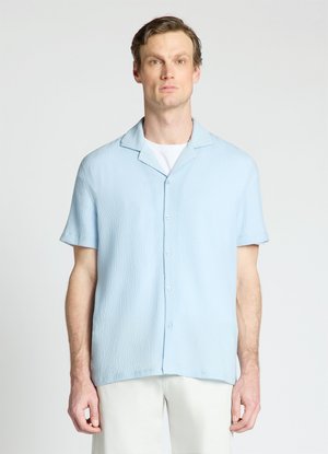 Uomo con camicia azzurra a maniche corte abbottonata sopra una T-shirt bianca, in piedi davanti a uno sfondo semplice.