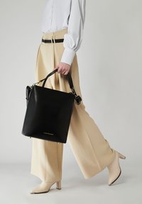 Svart läderhandväska med en strukturerad rektangulär form, med guldfärgade detaljer och ett avtagbart band, hålls av en person i beige byxor.