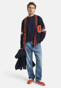 Cárdigan azul marino con rayas naranjas y la letra "G," combinado con una camiseta azul marino y jeans de color azul claro. Zapatillas naranjas y blancas completan el look.