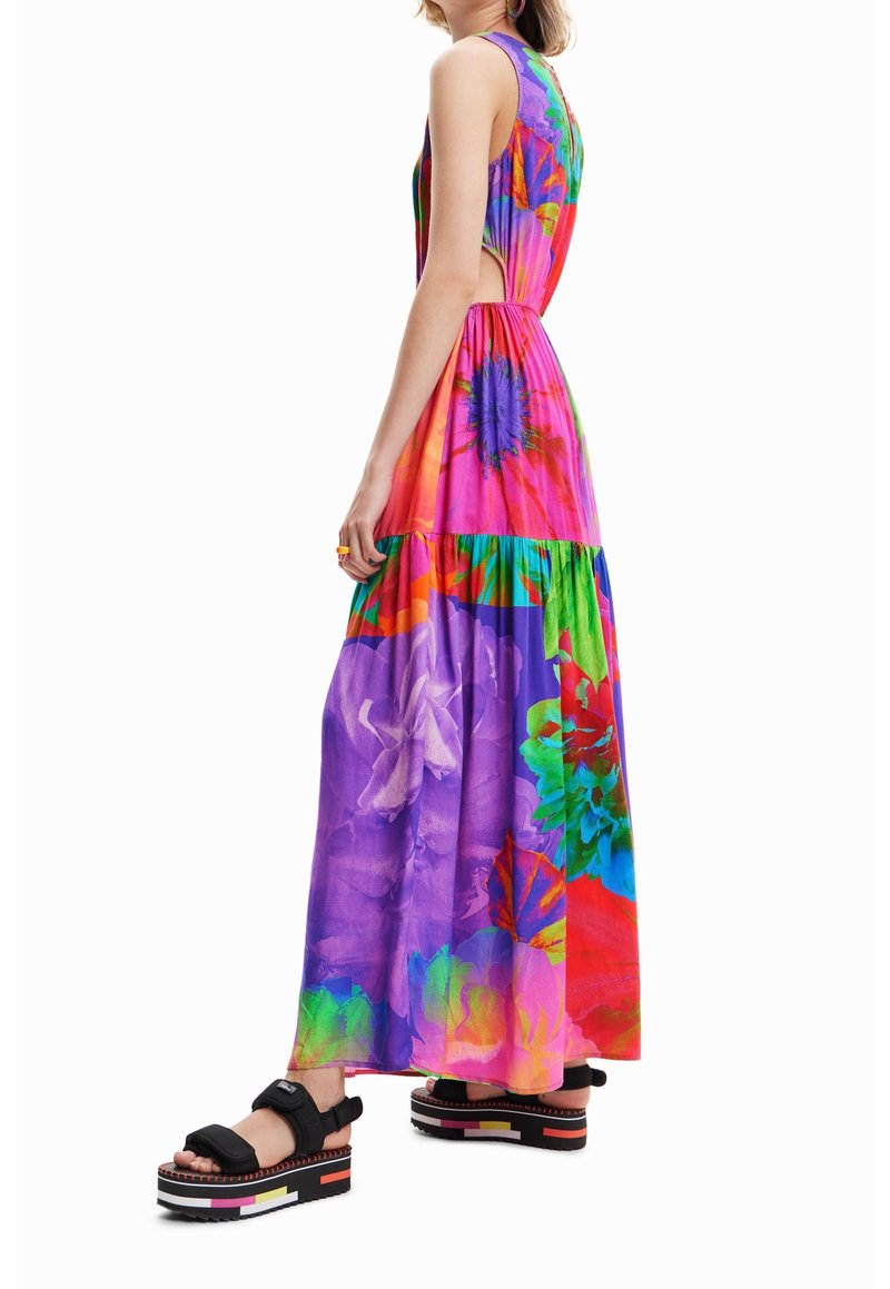 Robe maxi avec un imprimé floral vibrant en rose, violet, vert et bleu. Découpe sur le côté, design en couches, associée à des sandales à plateforme.