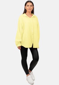 Camicia gialla con bottoni, realizzata in tessuto testurizzato, taglio oversize e colletto, abbinata a leggins neri e sneakers bianche.