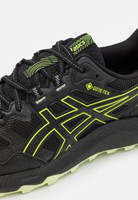Sapatilha de corrida preta com um upper texturizado, rótulo GORE-TEX e detalhes em verde neon. Sola AmpliFoam com padrão de pisada distinto.