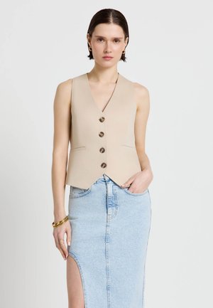 Donna che indossa un gilet beige senza maniche con bottoni e una gonna di jeans azzurro chiaro con spacco alto, con braccialetti e orecchini dorati, in piedi su sfondo bianco.