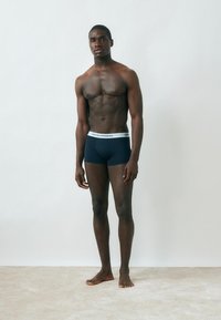 Atletyczny mężczyzna stoi boso przy gładkiej ścianie, nosi ciemnoniebieskie bokserki z białym paskiem z logo, patrzy przed siebie w luźnej pozie.