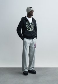 Μαύρο φούτερ με φερμουάρ και γραφικό σχέδιο, λευκό T-shirt, γκρι φόρμα με μωβ τύπωμα και μαύρα παπούτσια slip-on με μοτίβο. Γκρι σκουφί.