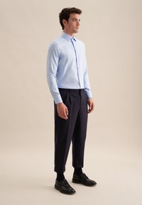 Uomo in piedi che indossa una camicia azzurra a maniche lunghe, pantaloni scuri plissettati, calzini neri e scarpe di pelle nere su uno sfondo beige.