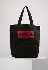 Svart canvas tote-väska med dubbla svarta handtag, med en stor röd Levi's-logotyp på framsidan och en stadig rektangulär form.