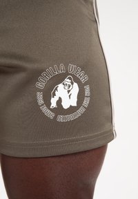 Olijfgroene sportshorts gemaakt van textuurstof, met een wit logo en tekst in een cirkelontwerp, voorzien van contrasterende zijstrepen.