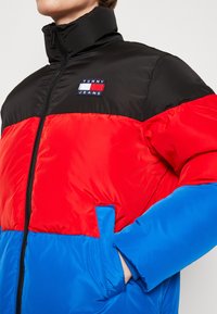 Giubbotto imbottito in nero, rosso e blu con colletto alto, chiusura con zip e patch del logo sul petto. Presenta tasche laterali.