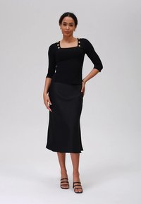 Musta istuva paita, jossa on kolme neljäsosaa hihaa ja koristeelliset kultaiset napit hartioissa, yhdistetty mustaan satiiniseen midihameeseen ja remmikenkiin.