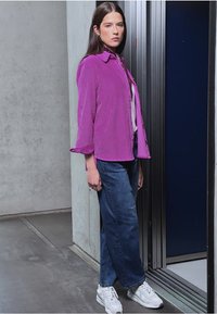 Magenta sztruksowa kurtka z kołnierzykiem, zestawiona z jasnofioletową bluzką i szerokimi granatowymi dżinsami. Białe trampki dopełniają strój.