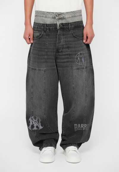 Jean en denim noir ample avec une ceinture élastique grise. Présente des graphiques argentés, y compris un logo et des éléments décoratifs sur les jambes.