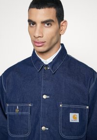 Jeansjacka i mörkblått med kontrasterande vit sömnad, utrustad med två bröstfickor, silverknappar och en Carhartt-logotyp.