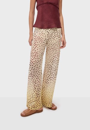 Femme portant un pantalon large beige à pois marron dégradant en jaune à l'ourlet, assorti à des sandales marron et un top sans manches bordeaux.