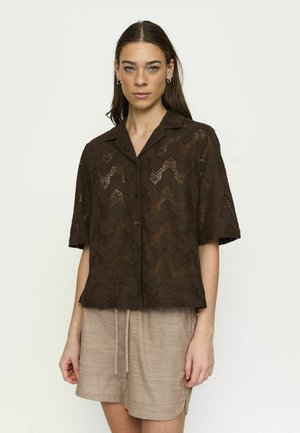 Nuori nainen, jolla on päällään tummanruskea, lyhythihainen pitsipaita ja beiget, kiristysnyörillä varustetut shortsit, seisoo vaaleaa taustaa vasten.