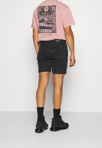 Rosa t-shirt med en svart grafisk design på baksidan, ihopparad med svarta denimshorts och svarta sportskor.