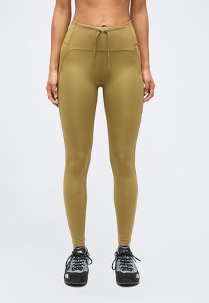 Γυναίκα που φοράει κολάν υψηλής μέσης σε ελιά πράσινο με τσέπες μπροστά και γκρι-μαύρα ορειβατικά παπούτσια, στέκεται μπροστά σε απλό φόντο.