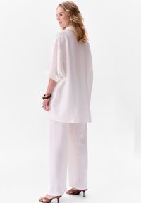 Witte oversized blouse met opgerolde mouwen, gecombineerd met witte wijde broek. Het gladde weefsel en de minimale details zorgen voor een schone, luchtige uitstraling.