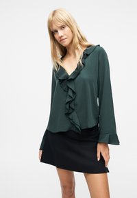 Νεαρή γυναίκα που φοράει σκούρο πράσινο μπλουζάκι με βολάν και μαύρη φούστα, στέκεται μπροστά σε απλό λευκό φόντο.