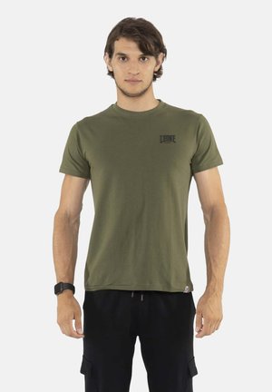Jeune homme aux cheveux courts portant un t-shirt vert olive et un pantalon noir, debout devant un fond clair uni.