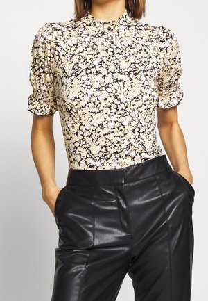 Vrouw die een korte mouw met bloemenprint draagt met opgeblazen mouwen en hooggetailleerde zwarte leren broek, handen in de zakken.