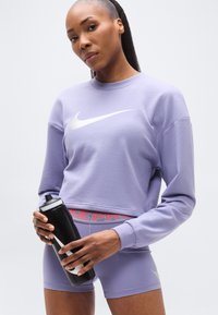 Μωβ cropped φούτερ με λευκό λογότυπο Nike, συνδυασμένο με μωβ σορτς που έχουν κόκκινη μέση με την επιγραφή "NIKE PRO". Κρατάει ένα μαύρο μπουκάλι νερού.